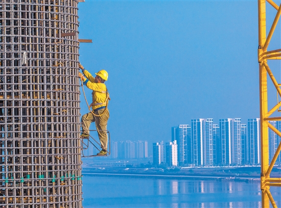 奮戰(zhàn)在百米索塔之上的建設(shè)者身影，與遠(yuǎn)處的高樓大廈遙呼相應(yīng)。