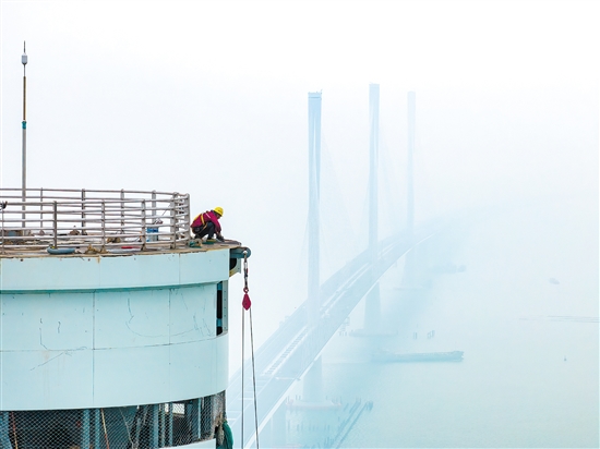 建設(shè)者在塔柱上施工，遠(yuǎn)處“海上長龍”若隱若現(xiàn)。